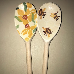 May include: Two wooden spoons with painted designs. One spoon features a floral pattern with yellow daffodils and green leaves. The other spoon has a pattern of brown bees on a white background.