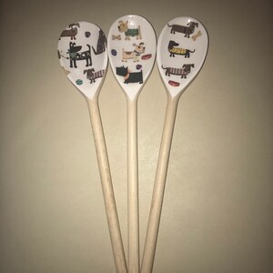 May include: Three wooden spoons with a white background and colorful dog illustrations. The spoons are decorated with various breeds of dogs, including dachshunds, terriers, and a black and white dog.