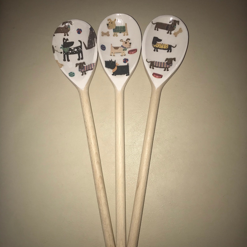 May include: Three wooden spoons with a white background and colorful dog illustrations. The spoons are decorated with various breeds of dogs, including dachshunds, terriers, and a black and white dog.