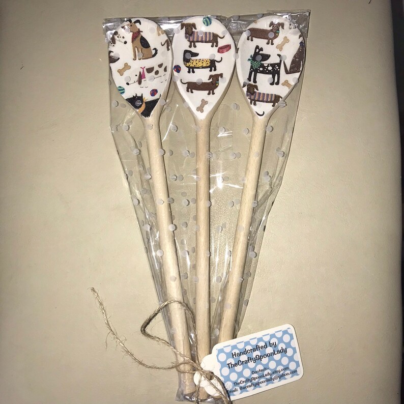 May include: Three wooden spoons with a white background and a dog pattern. The spoons are wrapped in clear plastic with white polka dots. A tag is attached to the spoons that reads "Handcrafted by TheCraftySpoonLady".