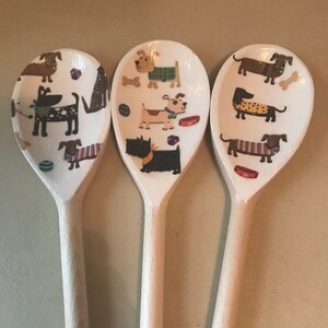 May include: Three wooden spoons with a white background and a pattern of dogs in various poses and colors. The dogs are wearing sweaters, collars, and bow ties.