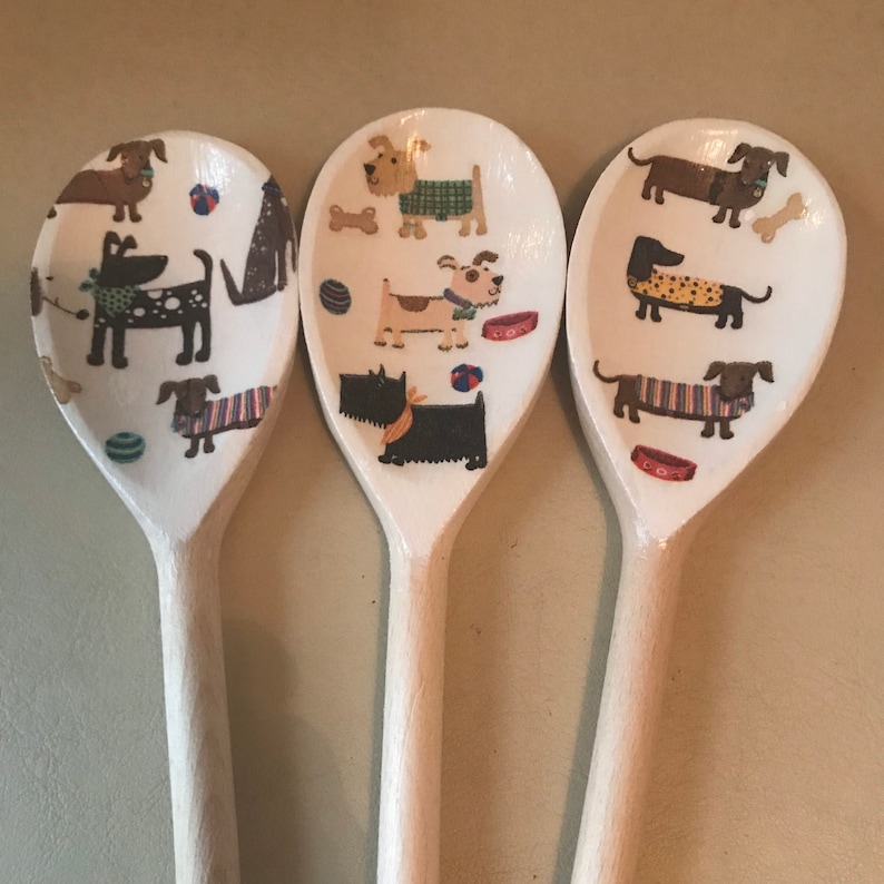 May include: Three wooden spoons with a white background and a pattern of dogs in various poses and colors. The dogs are wearing sweaters, collars, and bow ties.