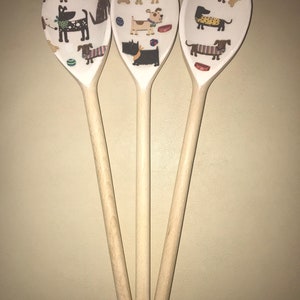 May include: Three wooden spoons with a white background and a pattern of dogs wearing clothes and playing with toys. The spoons are all the same size and shape, and they are arranged in a triangle.