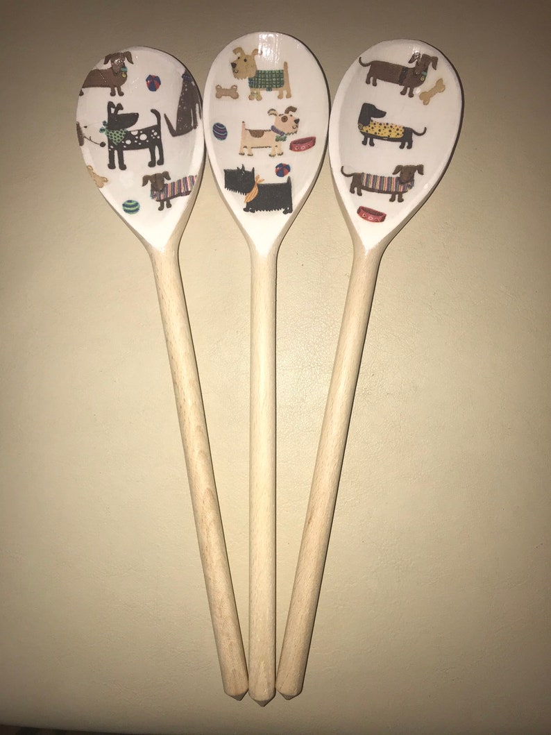 May include: Three wooden spoons with a white background and a pattern of dogs wearing clothes and playing with toys. The spoons are all the same size and shape, and they are arranged in a triangle.