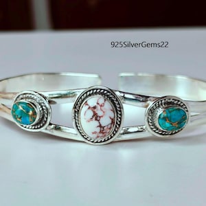 May include: A silver cuff bracelet with three oval-shaped stones. The central stone is white with brown veining, flanked by two turquoise stones. The bracelet has a polished finish and a simple, elegant design.