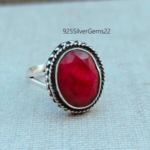 K&ouml;nnte beinhalten: Ein silberner Ring mit einem ovalen, facettierten, roten Edelstein. Der Edelstein ist von einem strukturierten, silbernen Rand umgeben. Der Ring teilt sich am Edelstein in zwei Teile. Der Text "925SilverGems22" ist im Hintergrund.