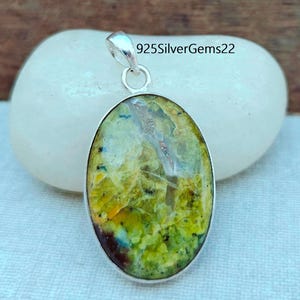 Peut inclure: Pendentif ovale avec une bélière argentée. Le pendentif présente une pierre précieuse vibrante et multicolore dans des tons de vert, de jaune et de marron. Le texte "925SilverGems22" est visible en haut à droite.