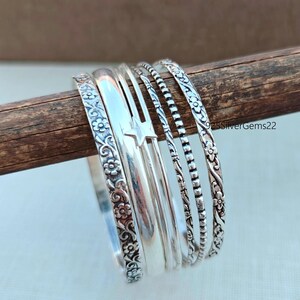 Peut inclure: Un ensemble de bracelets en argent, dont certains avec des motifs floraux et étoilés. Les bracelets sont présentés sur une cheville en bois, mettant en valeur leurs motifs détaillés et leurs textures variées. Les bracelets sont en argent.