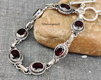May include: A silver bracelet with a toggle clasp features six red gemstone accents. The bracelet is on a brown rock surface. 925silvergems22 is printed on the rock.
