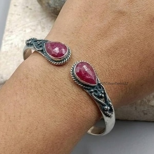 Puede incluir: Brazalete de plata con dos gemas rojas facetadas en forma de lágrima. El brazalete tiene un diseño abierto con detalles ornamentados en plata oscura alrededor de las gemas y en los brazos del brazalete. Las gemas están engastadas en un bisel de plata.