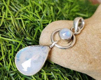 May include: A silver pendant with a teardrop-shaped moonstone and a smaller round moonstone set in a circle. The pendant is on a rock with green grass in the background.