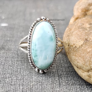 Op de afbeelding: Een zilveren ring met een lichtblauwe ovale edelsteen. De ring heeft een touwachtig ontwerp rond de rand van de steen en kleine kralen rond de zetting.