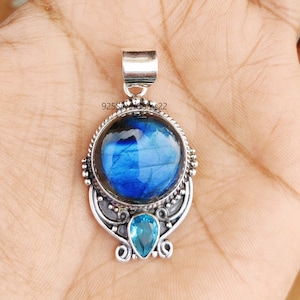 Puede incluir: Un colgante de plata con una piedra preciosa de labradorita azul y una piedra preciosa de topacio azul más pequeña. El colgante tiene un diseño intrincado con una argolla de plata.