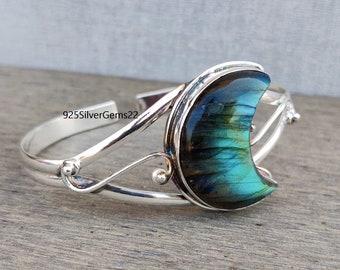 May include: Silver crescent moon bracelet with a blue-green labradorite gemstone. The bracelet has a delicate, ornate design.