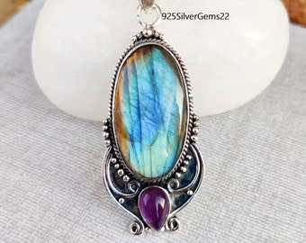 May include: Silver pendant featuring a large oval labradorite gemstone with iridescent blue and green hues, set in a silver filigree design with a smaller amethyst gemstone at the bottom. 925SilverGems22 is printed on the top of the pendant.