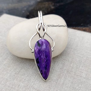 Puede incluir: Un colgante de plata con una piedra púrpura y negra. El colgante tiene forma de lágrima y presenta un diseño delicado e intrincado. La piedra está engastada en un engaste de plata y tiene un patrón único y arremolinado.