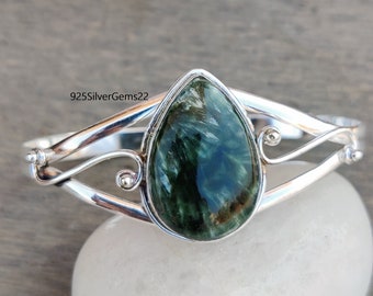 May include: A silver bangle bracelet featuring a green aventurine gemstone. The gemstone is teardrop shaped and is set in a silver bezel. The bracelet has a delicate, twisted design.
