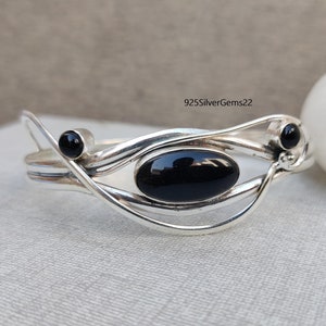 Może przedstawiać: Srebrna bransoletka z zapięciem typu bangle, ozdobiona czarnym owalnym kamieniem szlachetnym i dwoma mniejszymi czarnymi okrągłymi kamieniami szlachetnymi. Bransoletka ma delikatny i skomplikowany wzór.
