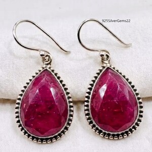 May include: A pair of silver teardrop earrings with large, faceted, ruby-red gemstones. The gemstones are surrounded by a beaded silver border. The earrings have a simple hook design. The text "925SilverGems22" is visible in the background.