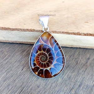 May include: A silver pendant featuring a teardrop-shaped ammonite fossil with a brown and blue iridescent shell. The pendant is on a wooden surface.