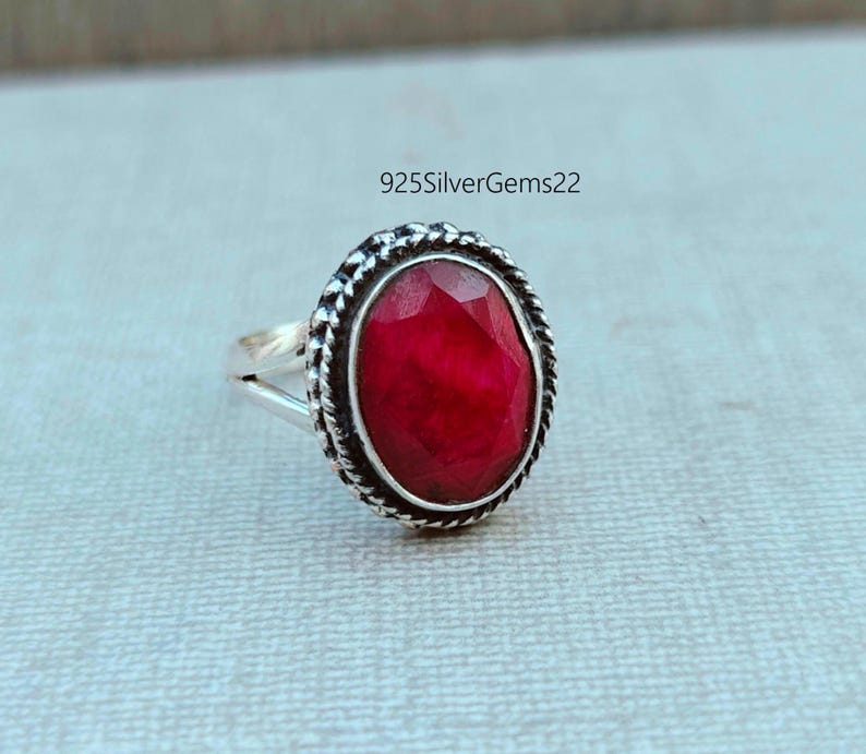 K&ouml;nnte beinhalten: Ein silberner Ring mit einem ovalen, facettierten, roten Edelstein. Der Edelstein ist von einem strukturierten, silbernen Rand umgeben. Der Ring teilt sich am Edelstein in zwei Teile. Der Text "925SilverGems22" ist im Hintergrund.