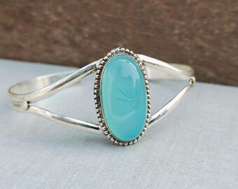 May include: A silver cuff bracelet featuring a large, oval, light blue gemstone. The gemstone is surrounded by a beaded border and set in a silver band. The bracelet has a simple, elegant design.