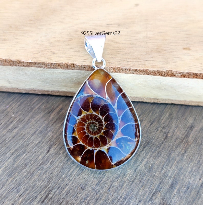 May include: A silver pendant featuring a teardrop-shaped ammonite fossil with a brown and white spiral pattern. The pendant is on a wooden surface.
