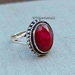 K&ouml;nnte beinhalten: Ein silberner Ring mit einem gro&szlig;en, ovalen, tiefroten Edelstein. Der Edelstein ist in einer dekorativen L&uuml;nette mit strukturiertem Rand gefasst. Das Ringband ist oben geteilt. Der Text "925SilverGems22" ist sichtbar.