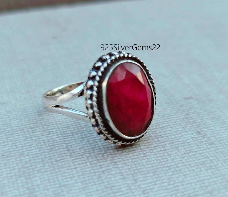 K&ouml;nnte beinhalten: Ein silberner Ring mit einem gro&szlig;en, ovalen, tiefroten Edelstein. Der Edelstein ist in einer dekorativen L&uuml;nette mit strukturiertem Rand gefasst. Das Ringband ist oben geteilt. Der Text "925SilverGems22" ist sichtbar.