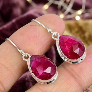 May include: A pair of silver drop earrings featuring large, faceted, teardrop-shaped, ruby-red gemstones. The earrings have a simple, elegant design with a polished finish. The text "925SilverGems22" is visible.