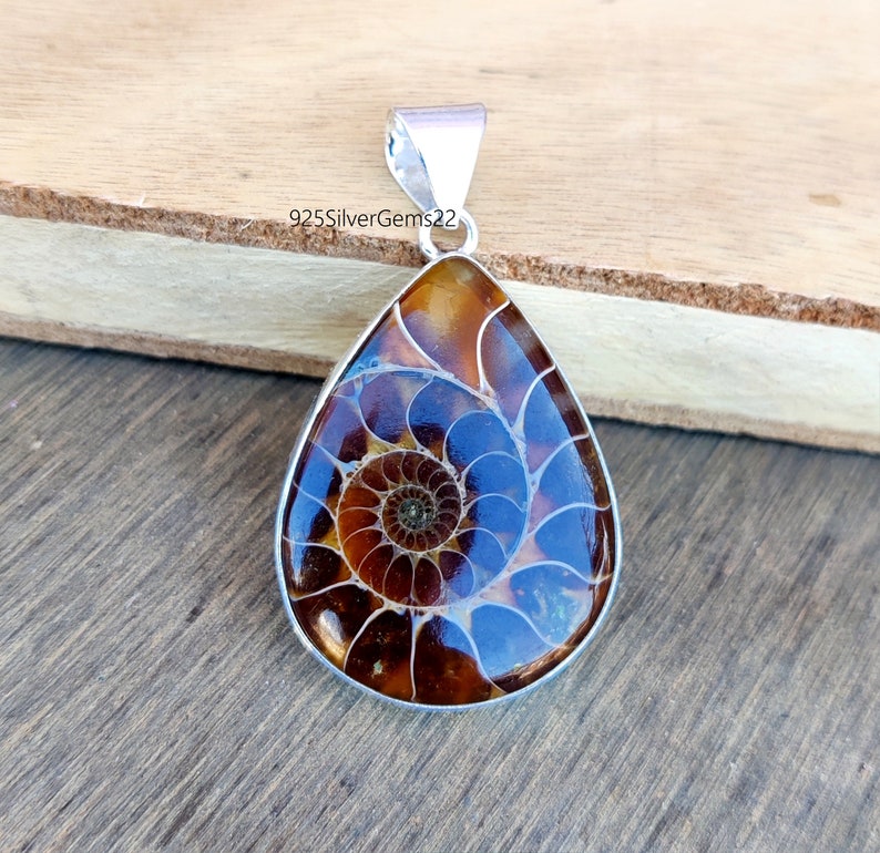 May include: A silver pendant featuring a brown and blue ammonite fossil with a spiral pattern. The pendant is in the shape of a teardrop and has a silver bail.