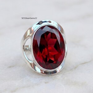 May include: A silver ring featuring a large, oval-cut, deep red gemstone. The ring has a polished, rounded band with openwork details on the sides. The gemstone is the focal point, showcasing a faceted surface that catches the light.