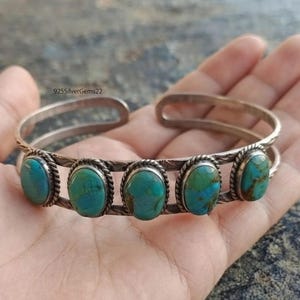 Könnte beinhalten: Ein silbernes Armband mit fünf ovalen Türkissteinen mit braunen Matrixmustern. Die Steine sind in gedrehte Silberdrahtfassungen eingefasst und das Band besteht aus zwei parallelen Silberdrähten. Der Text 925SilverGems22 ist sichtbar.