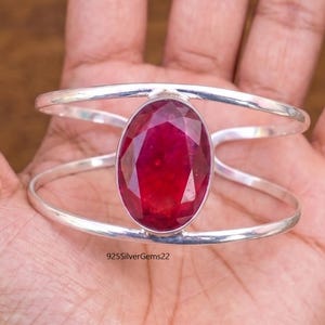May include: A silver cuff bracelet featuring a large, oval-cut ruby gemstone. The ruby is a deep red color and is set in a polished silver band. The bracelet has a modern, open design.