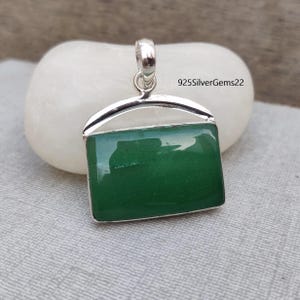 Puede incluir: Un colgante de plata con una piedra preciosa verde. El colgante tiene forma rectangular con esquinas redondeadas. La piedra preciosa está engastada en un marco de plata. El colgante cuelga de una cadena de plata.