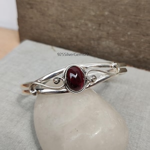 Pode incluir: Um bracelete de prata com um fecho circular e uma pedra vermelha engastada em um delicado design em espiral. 925SilverGems22 está impresso no fundo.
