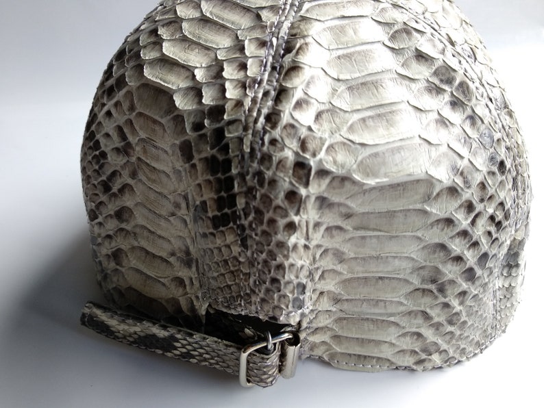 May include: A grey snakeskin cap with a silver buckle closure. The cap is made of genuine snakeskin and has a unique texture.