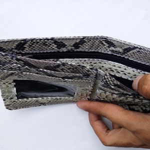 May include: A brown and white snakeskin wallet with a zipper closure and a clear plastic window. The wallet is open and a hand is holding it.