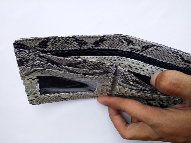 May include: A brown and white snakeskin wallet with a zipper closure and a clear plastic window. The wallet is open and a hand is holding it.
