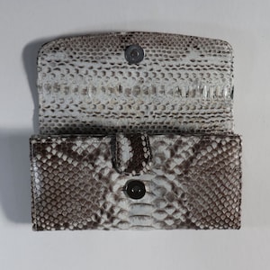 May include: Open, rectangular wallet with a snakeskin pattern in shades of white, grey, and brown. The wallet has a magnetic snap closure and a flap that opens to reveal the interior. The wallet is made of a textured material.