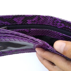 May include: Purple snakeskin wallet with a zipper closure and a clear plastic window. The wallet is open and a hand is reaching inside.