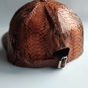 May include: A brown leather baseball cap with a snakeskin pattern. The cap has a strap with a metal buckle.