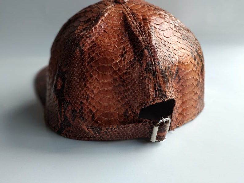 May include: A brown leather baseball cap with a snakeskin pattern. The cap has a strap with a metal buckle.