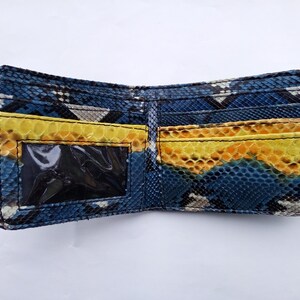 May include: Open bi-fold wallet with a snakeskin pattern in shades of blue, yellow, and gray. The interior features yellow card slots and a clear ID window. The wallet is made of a textured material.