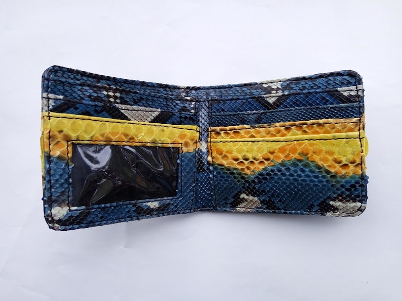 May include: Open bi-fold wallet with a snakeskin pattern in shades of blue, yellow, and gray. The interior features yellow card slots and a clear ID window. The wallet is made of a textured material.