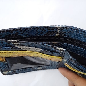May include: Open view of a bi-fold wallet made of blue, black, and yellow snakeskin. The wallet has a black zipper closure and a clear ID window. The interior is black, and the wallet is open, revealing the compartments.