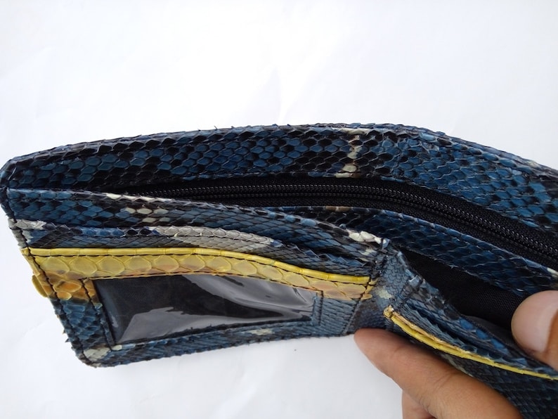 May include: Open view of a bi-fold wallet made of blue, black, and yellow snakeskin. The wallet has a black zipper closure and a clear ID window. The interior is black, and the wallet is open, revealing the compartments.