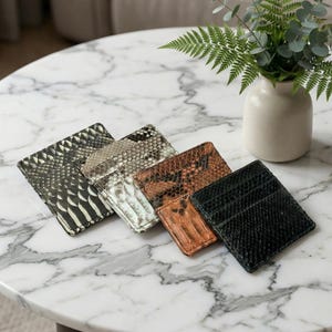 May include: Four rectangular card holders with textured snake-skin patterns in black, metallic silver, brown, and solid black. Each item features multiple slots for organizing cards, arranged in a row on a white marble surface.