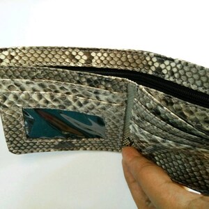 May include: Open bi-fold wallet with a snakeskin pattern in shades of gray, brown, and black. The wallet has multiple card slots, a clear ID window, and a zippered compartment. The interior is a lighter gray color.