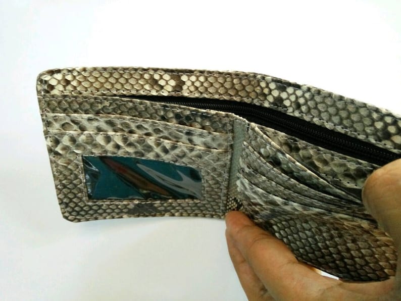 May include: Open bi-fold wallet with a snakeskin pattern in shades of gray, brown, and black. The wallet has multiple card slots, a clear ID window, and a zippered compartment. The interior is a lighter gray color.